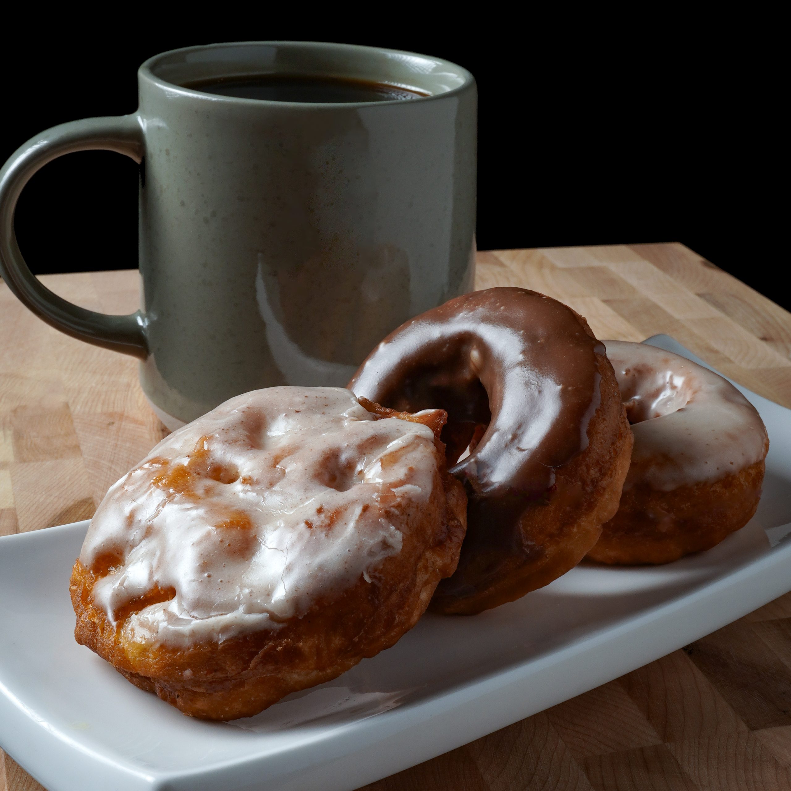 3 different donuts on a white plate with a steaming coffee cup in the background.